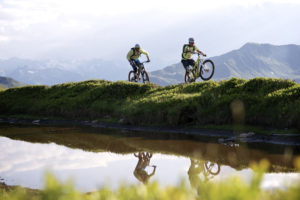 zwei Montainbiker spiegeln sich auf einem Hügel im darunter befindlichen Wasser